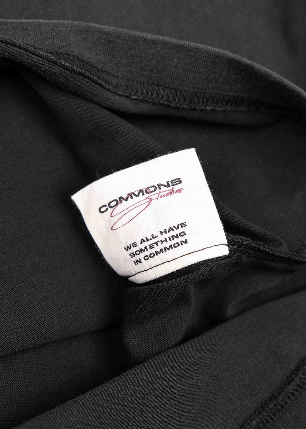 Close-up of a black knit T-shirt with visible stitching and a white Commons Studio label with black and red lettering.