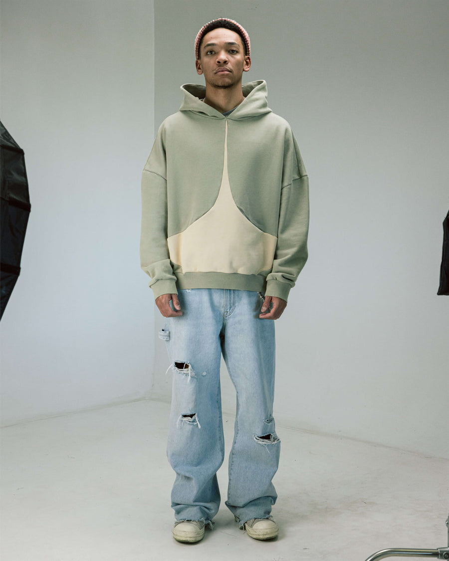 Model wearing a sage green hoodie with a cream front panel, ripped light blue jeans, and a knit beanie in a studio.