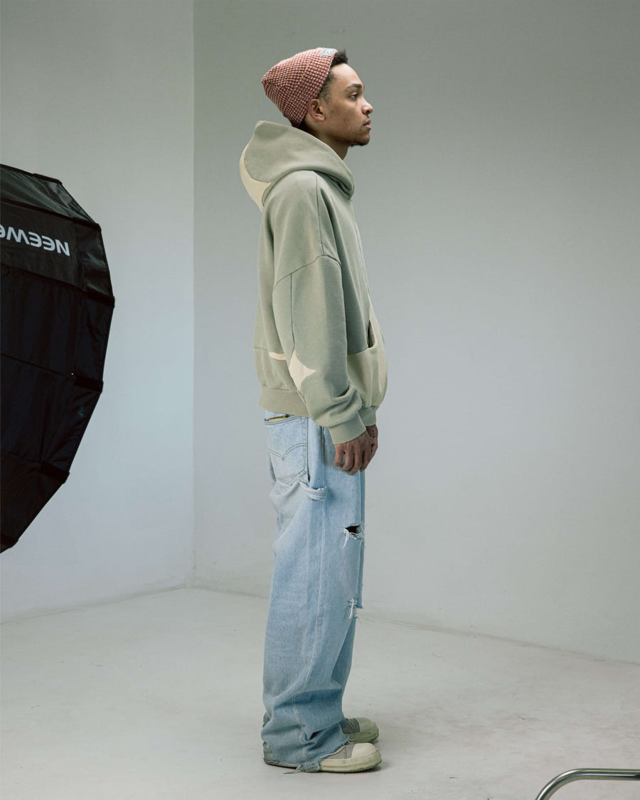 Model in a sage green hoodie with cream star details, distressed light blue jeans and a red knit beanie in a studio.
