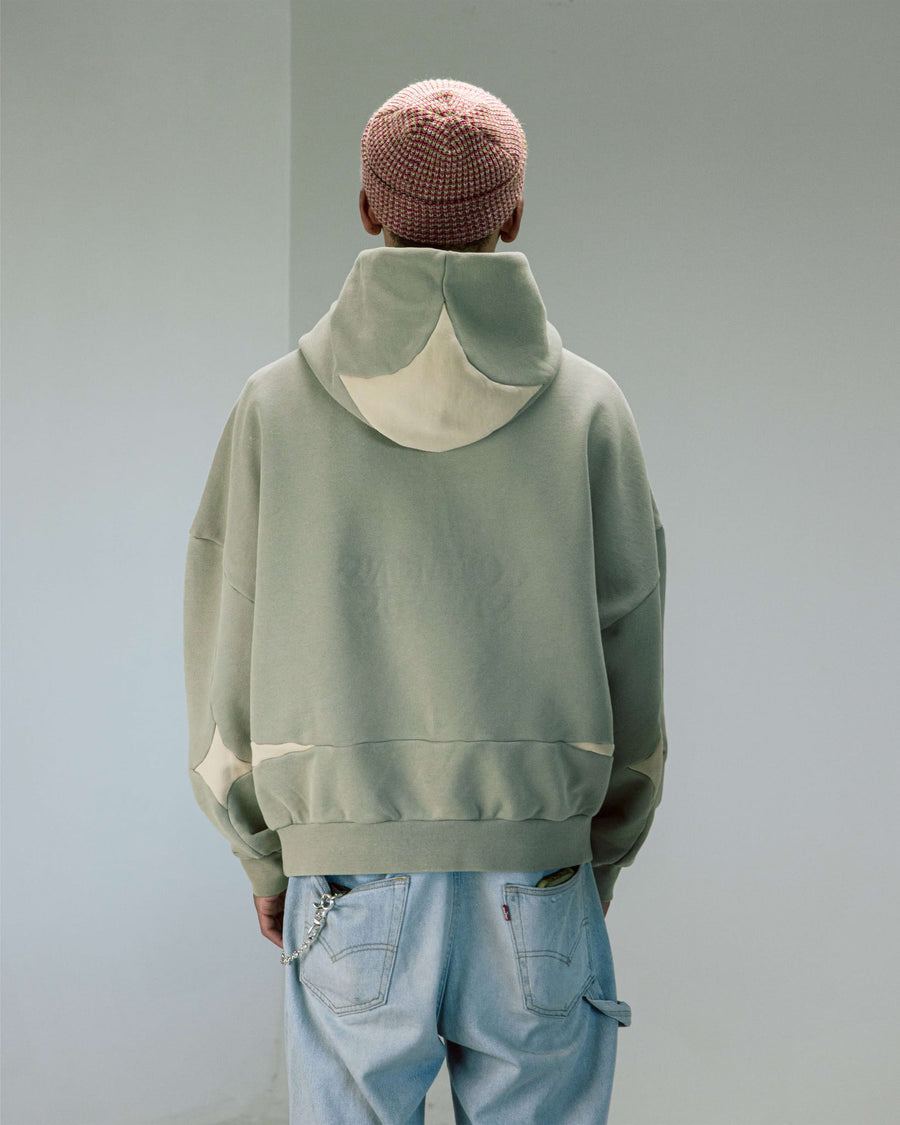 Back view of a person wearing a sage green hoodie with cream star details, light blue jeans, and a multicolored knit cap.