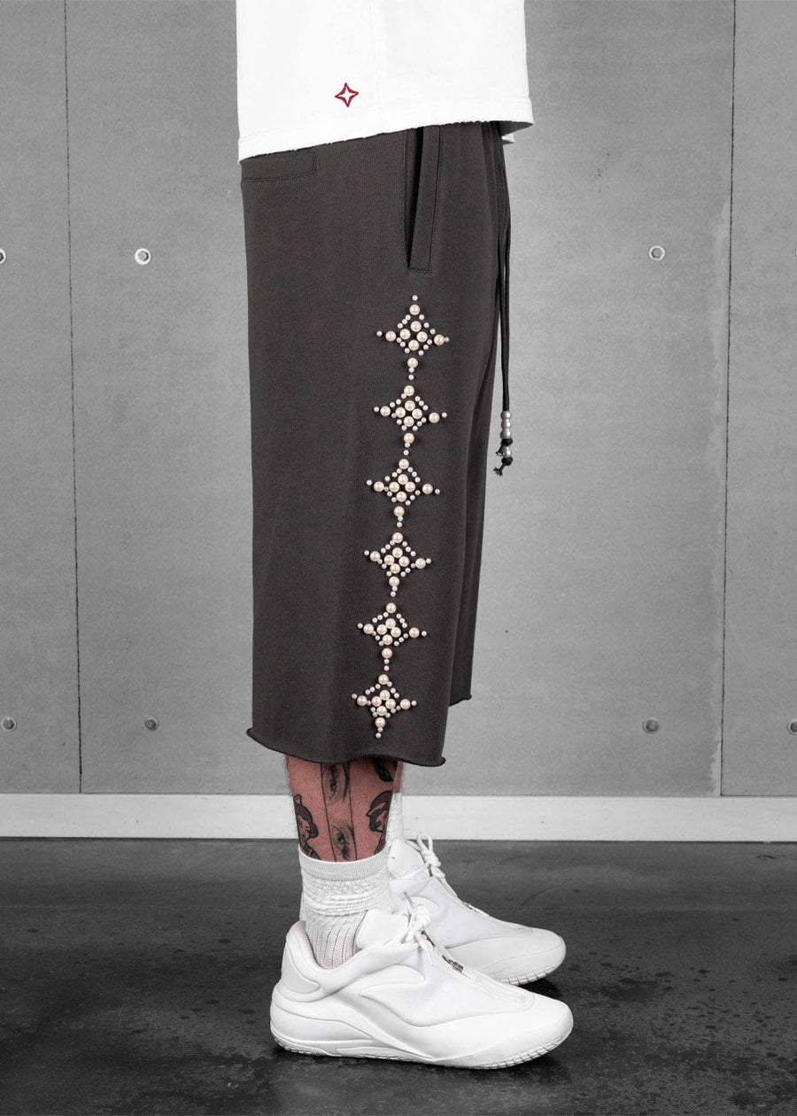 Model wearing dark taupe-gray long shorts with pearl-like star embellishments, raw hems, and white sneakers.