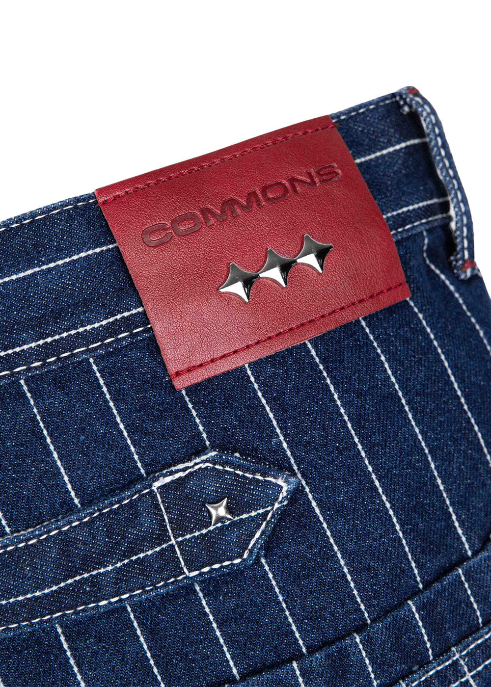 Close-up of blue pinstriped denim pants with a red COMMONS leather patch, silver studs, and white stitching.