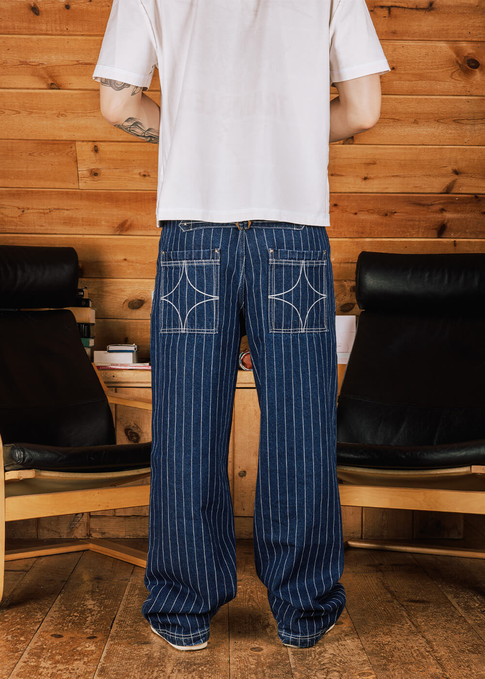 Back view of blue pinstriped denim pants with decorative rear pocket stitching, worn with a white T-shirt in a wood-paneled room.