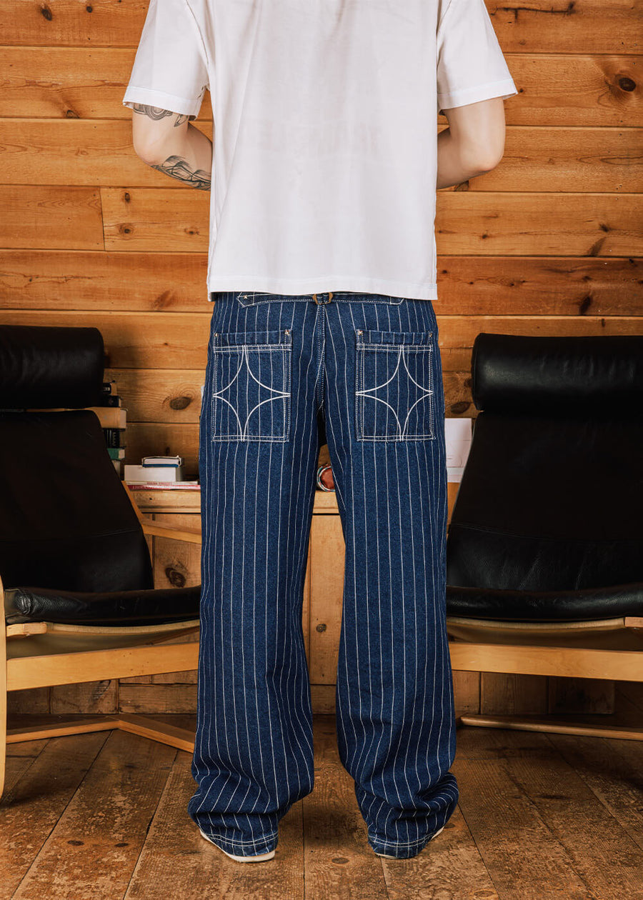 Back view of blue pinstriped denim pants with decorative rear pocket stitching, worn with a white T-shirt in a wood-paneled room.