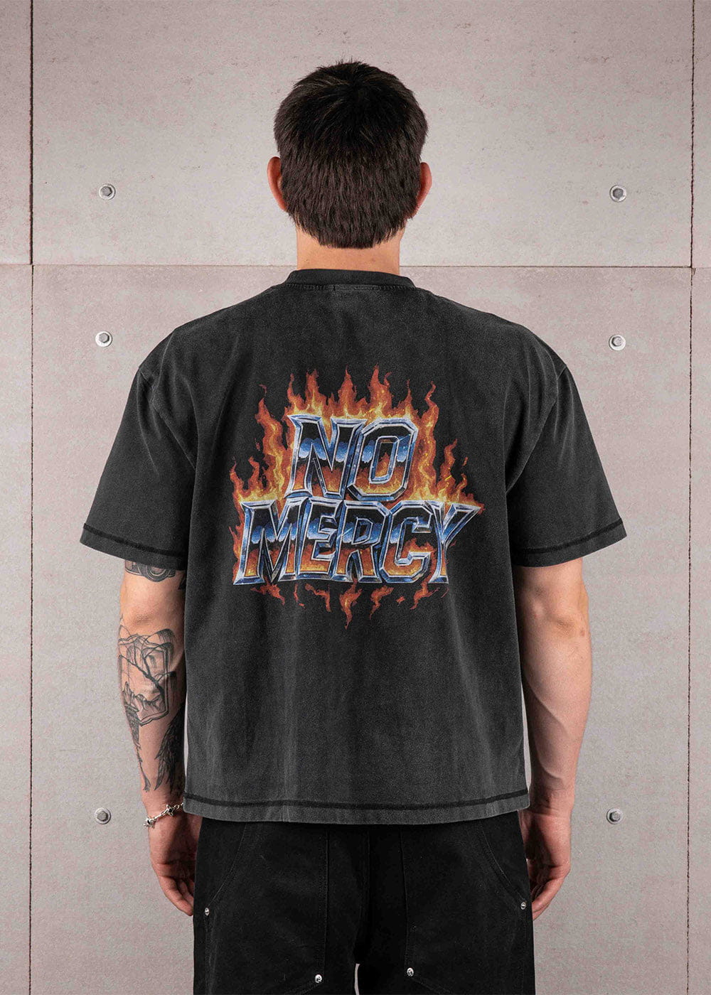 Back view of a person wearing a washed black T-shirt with NO MERCY lettering and orange flames against a concrete wall.