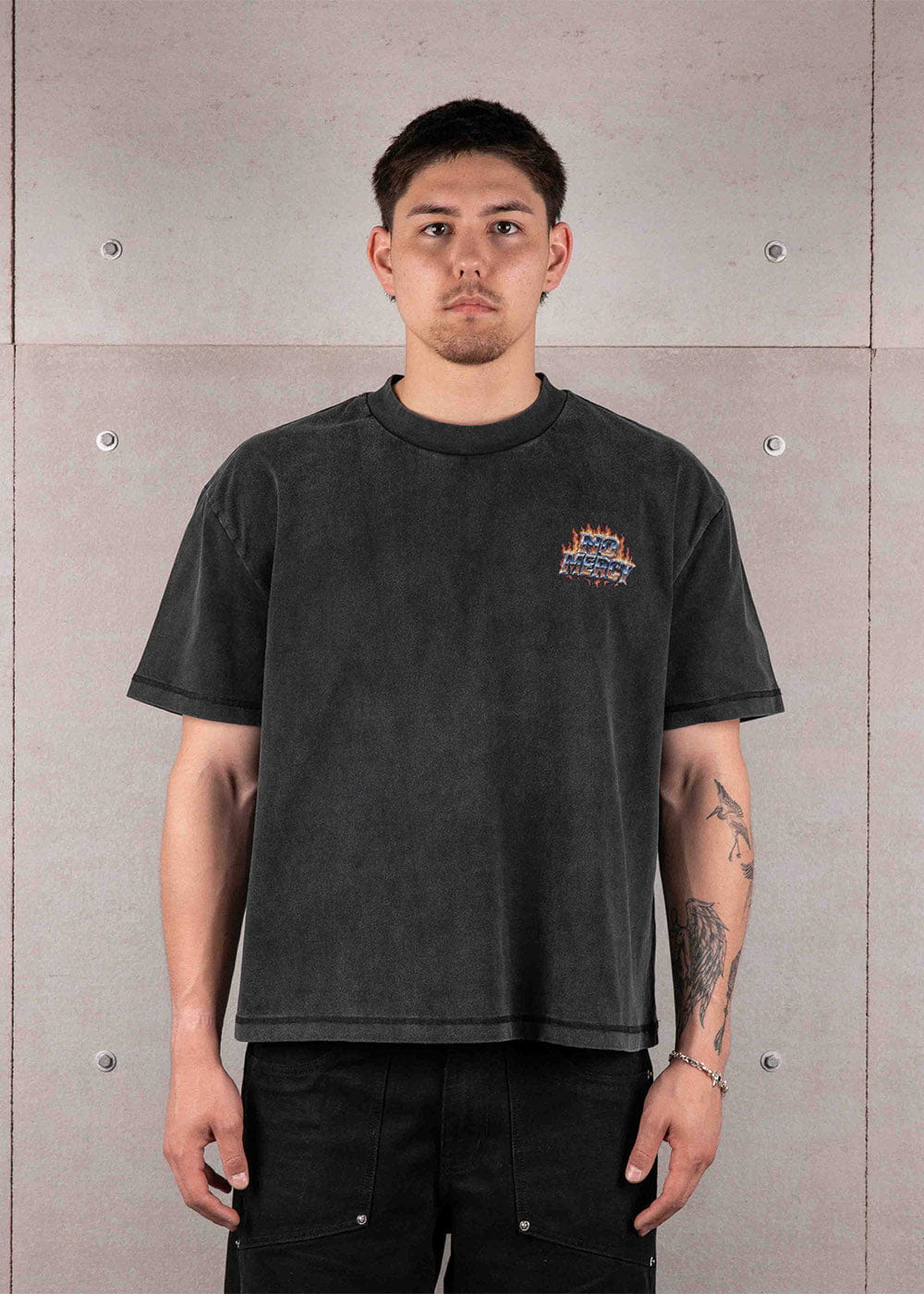 Man wearing a washed black crewneck T-shirt with a small flame chest graphic and black pants against a gray concrete wall.