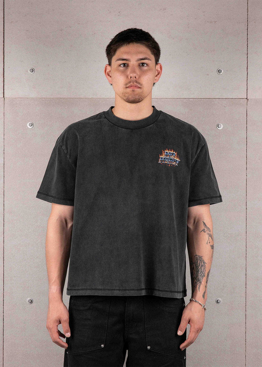 Man wearing a washed black crewneck T-shirt with a small flame chest graphic and black pants against a gray concrete wall.