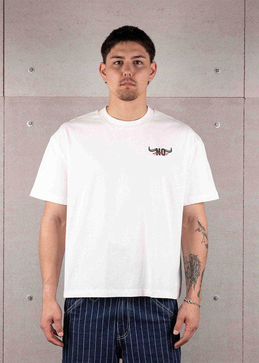 Man wearing a white crewneck T-shirt with a small black and red chest logo and navy striped pants against a concrete wall.