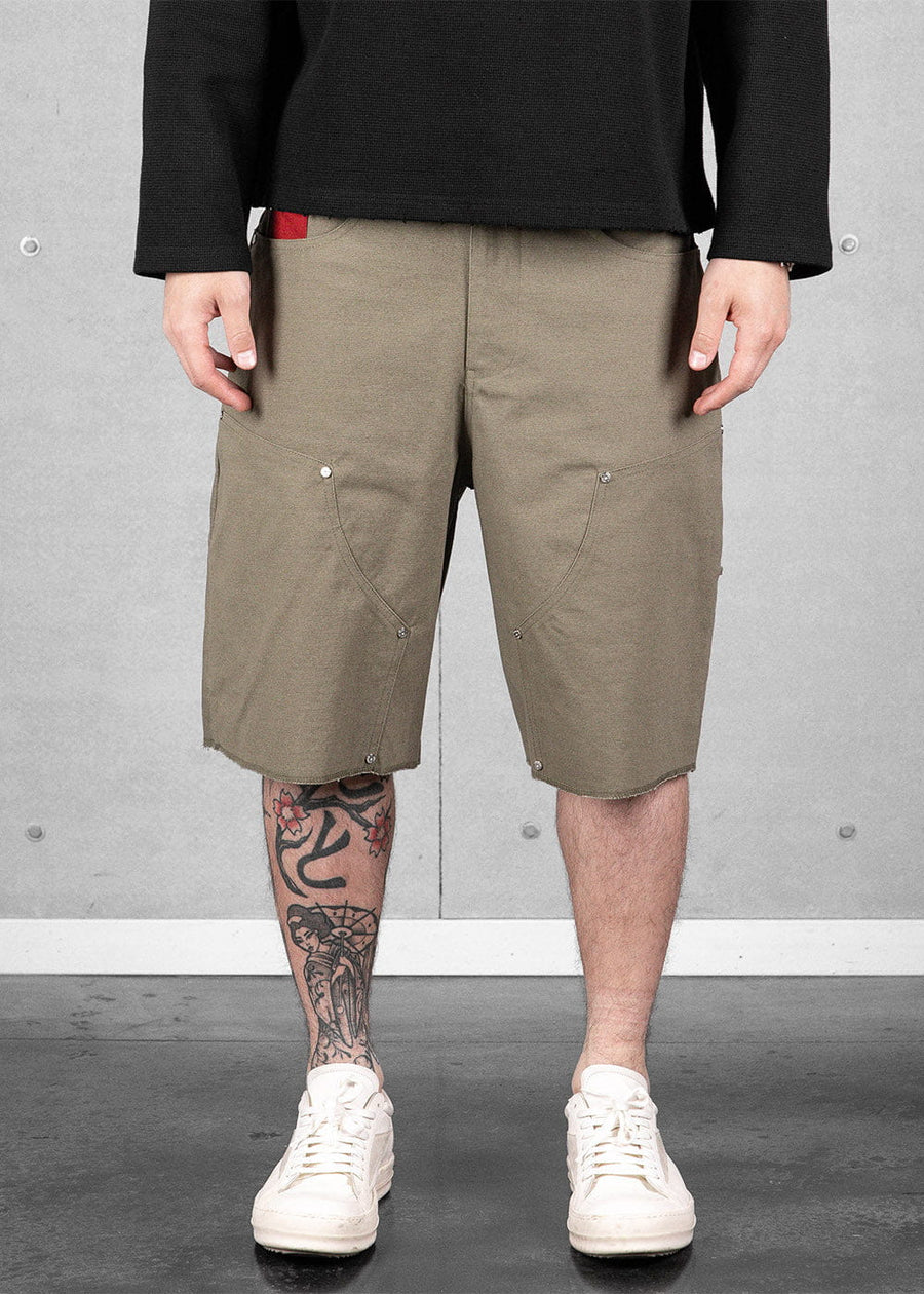 Olive green canvas knee-length shorts with curved seams and metal rivets, worn with a black top and white sneakers.
