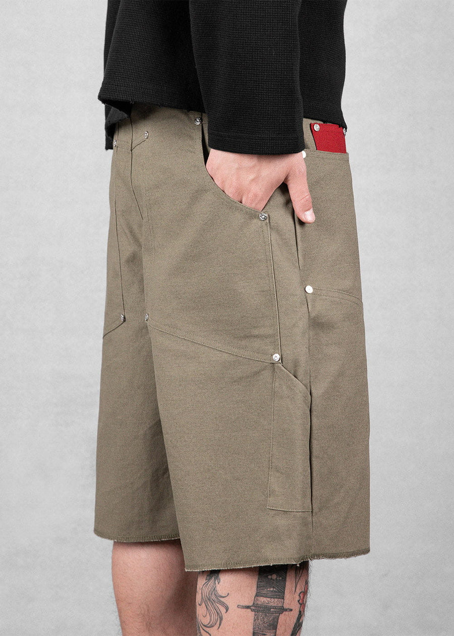 Olive canvas shorts with large riveted pockets and raw hems, worn with a black top against a gray background.