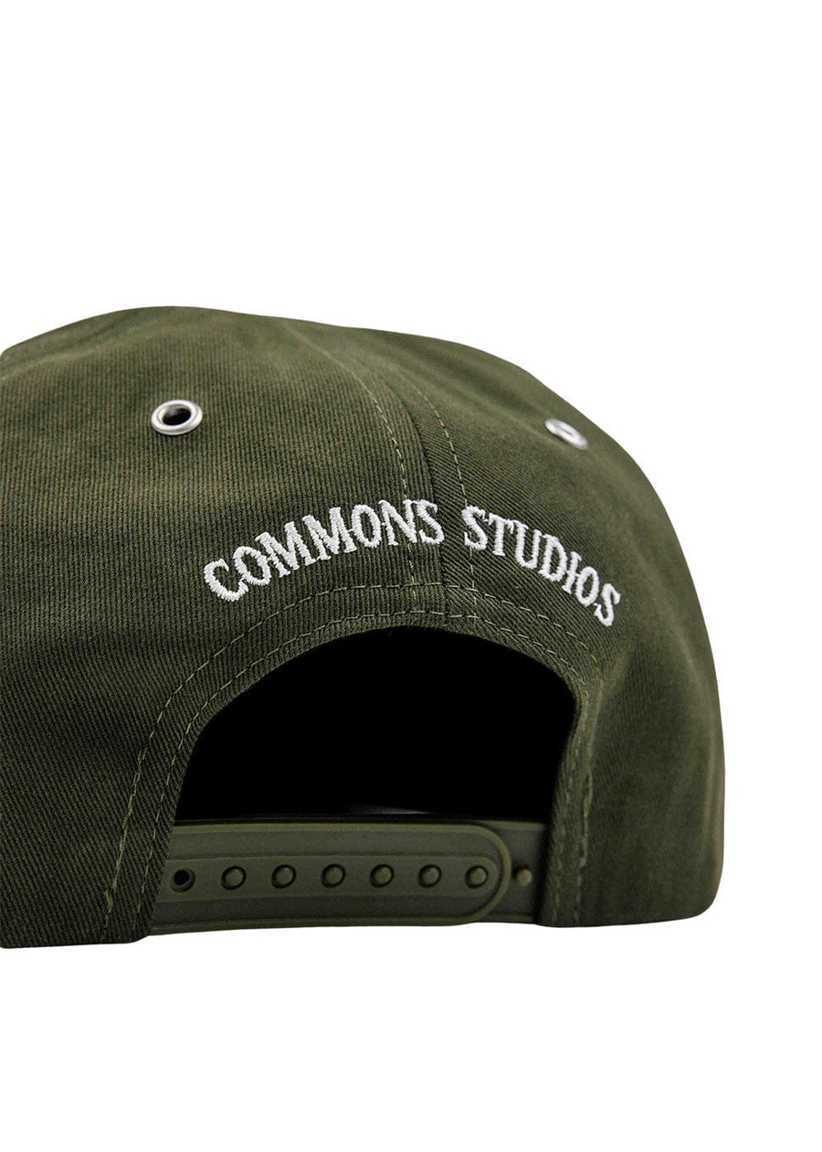 Back of an olive green snapback cap with white COMMONS STUDIOS embroidery, metal eyelets, and an adjustable strap.