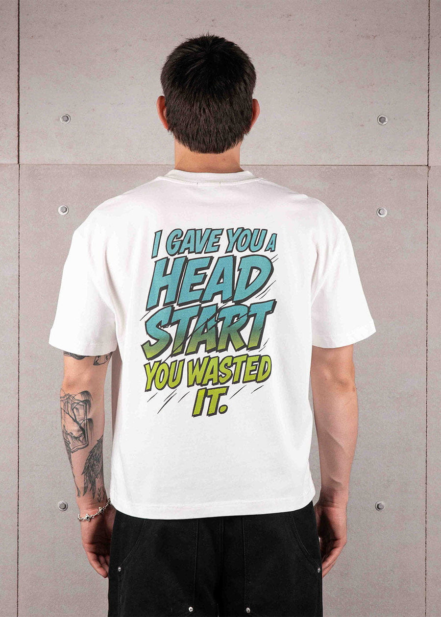 Person facing a concrete wall wearing a white T-shirt with large teal and lime text on the back and black pants.
