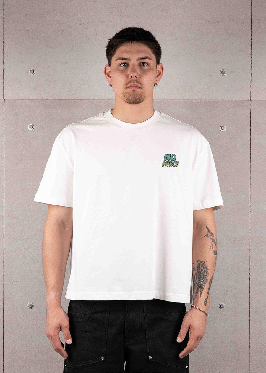 Man wearing a white short-sleeve T-shirt with a small teal and lime NO MERCY chest graphic against a gray wall.