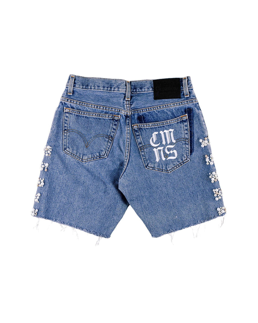 Back view of blue denim shorts with raw hems, pearl-like side embellishments, and white lettering on one back pocket.