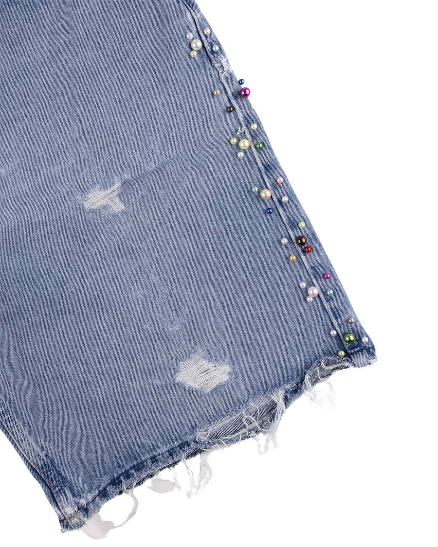 Close-up of light blue denim with distressed patches, a frayed hem, and multicolored pearl-like beads along the side seam.