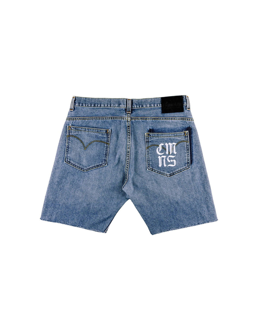 Back view of blue denim shorts with raw hems, two pockets, white lettering on one pocket, and a black waistband patch.