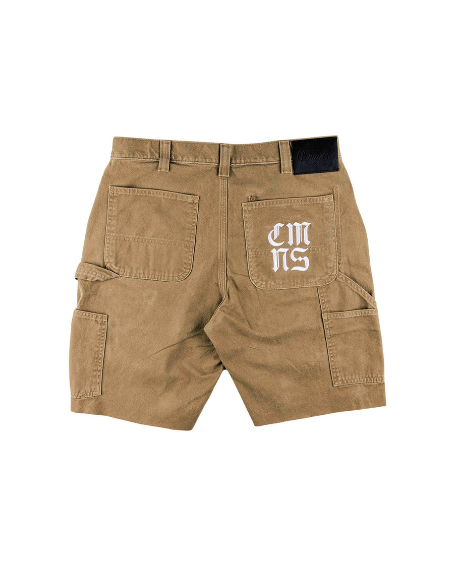 Back view of tan carpenter shorts with utility pockets, hammer loop, white pocket lettering, and a black waistband patch.