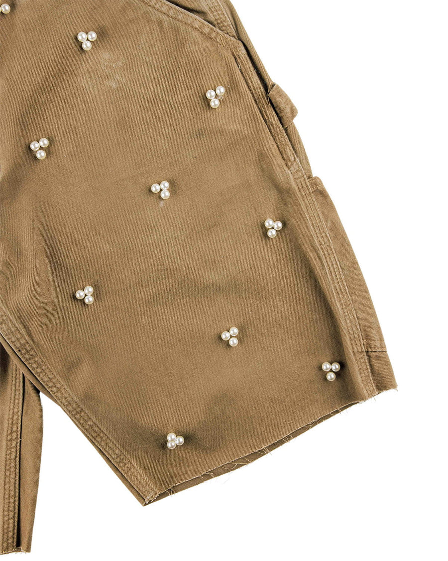 Close-up of tan twill shorts with white pearl bead clusters, stitched seams, and raw hems on a white background.