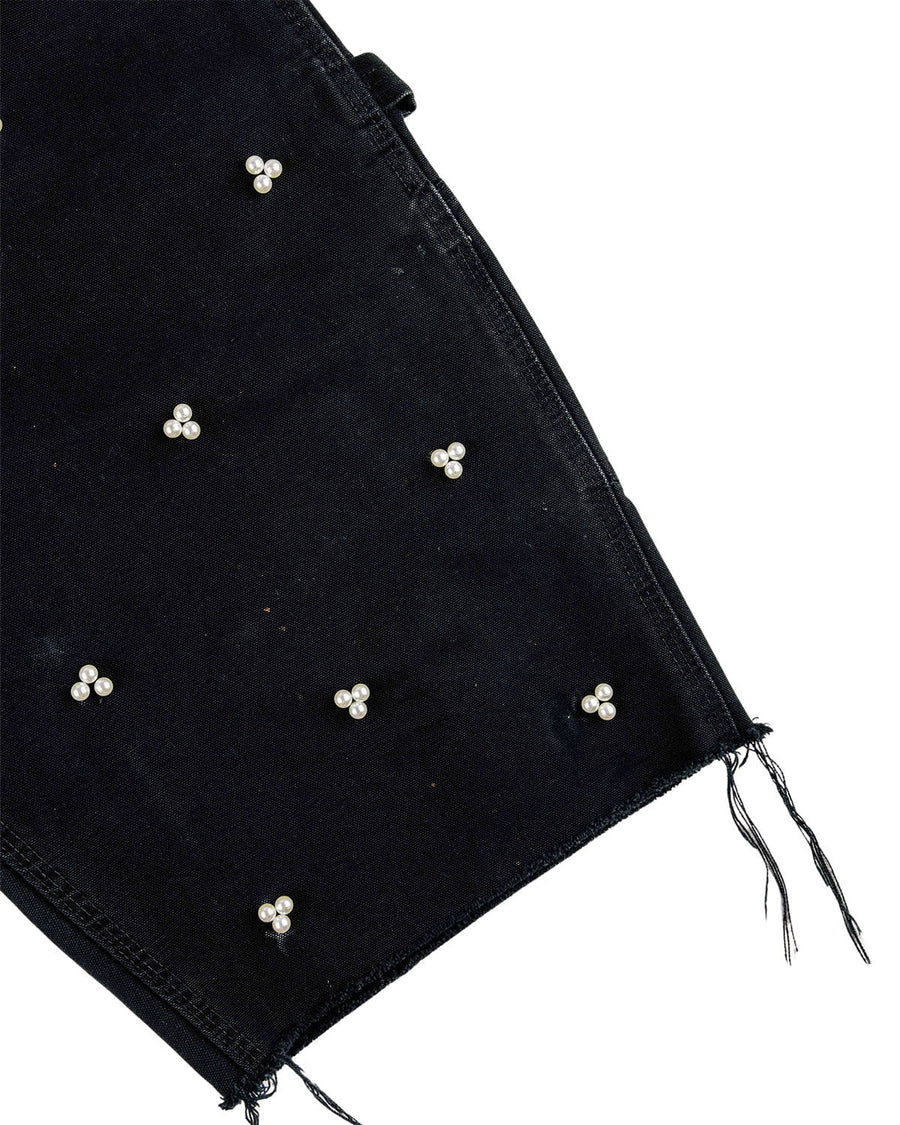 Close-up of black denim with clusters of three white pearl beads and a frayed raw hem on a white background.
