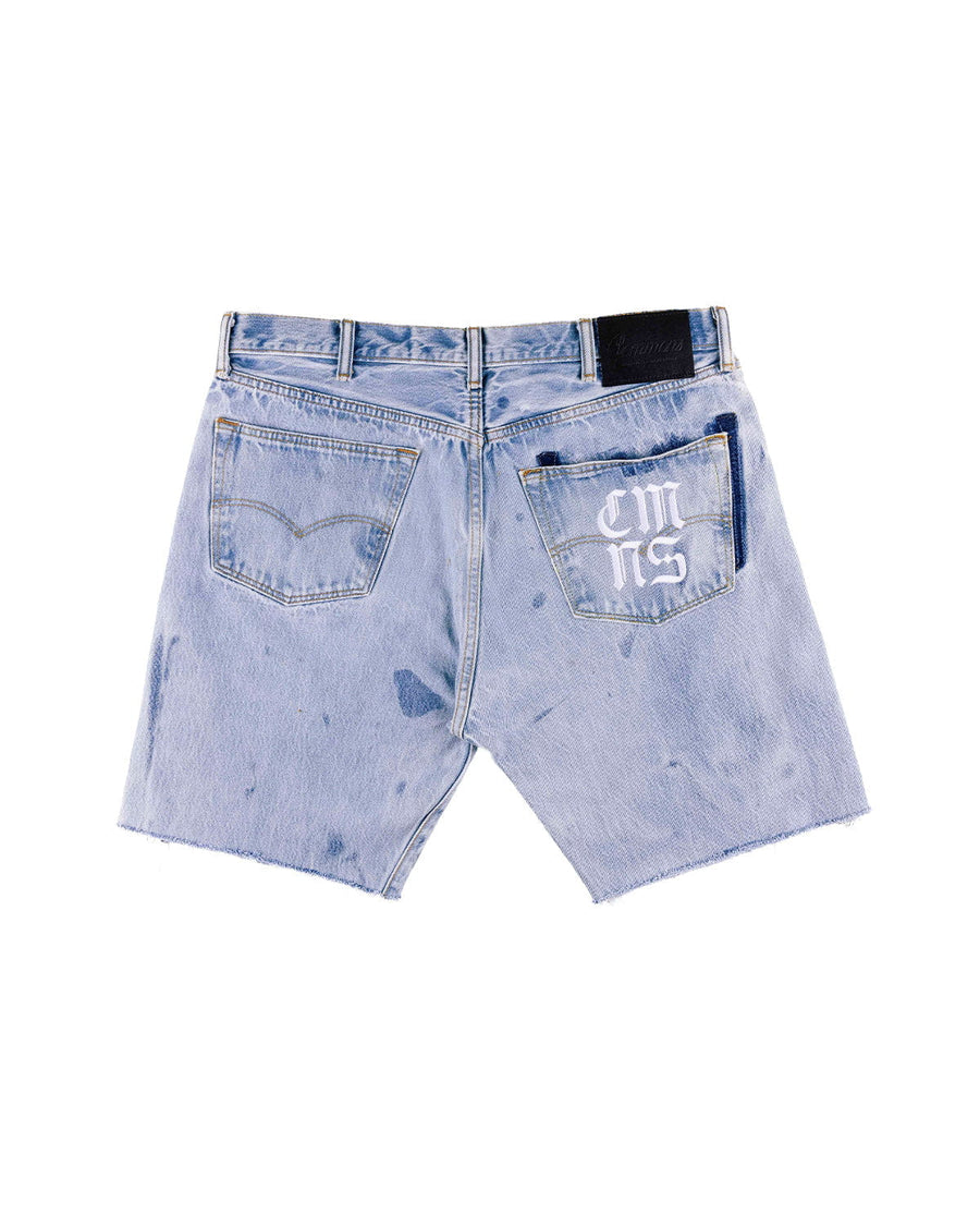 Back view of light blue denim shorts with raw hems, dark faded patches, and white lettering on one back pocket.
