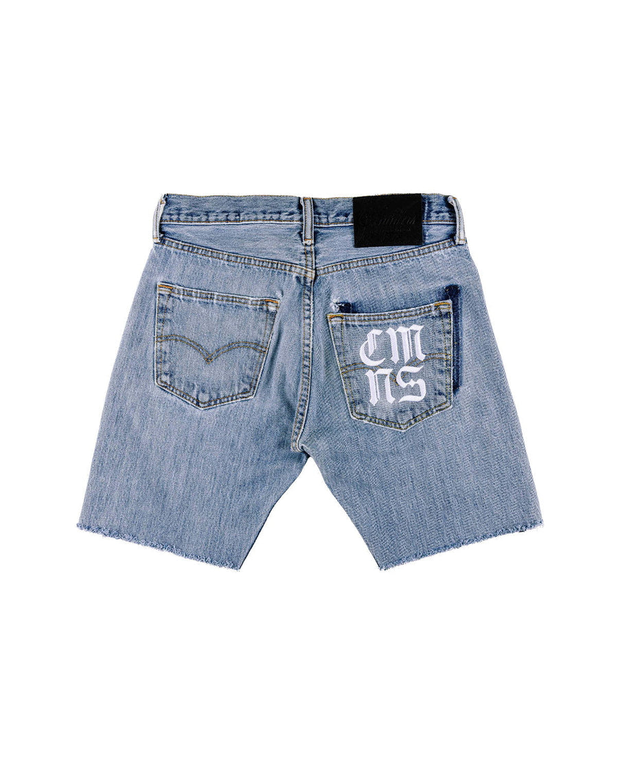 Back view of light blue denim shorts with raw hems, two pockets, white lettering on one pocket, and a black waistband patch.