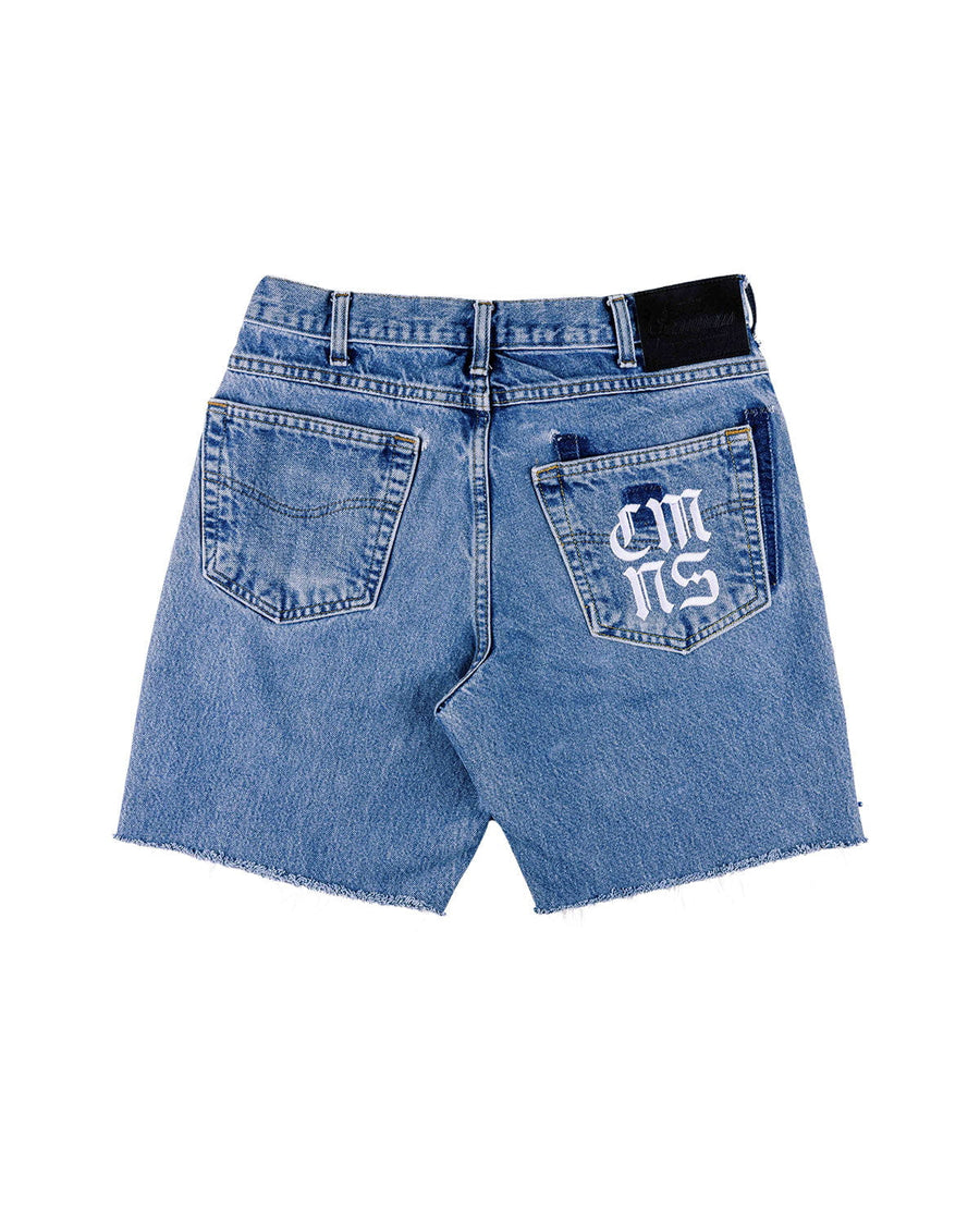 Back view of blue denim shorts with raw hems, two pockets, white Gothic lettering, and a black waistband patch.