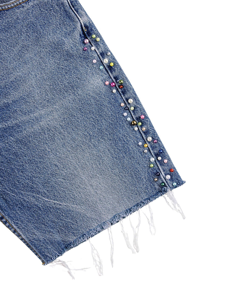 Close-up of blue denim shorts with multicolored pearl-like beads along the side seam and a frayed hem on white.