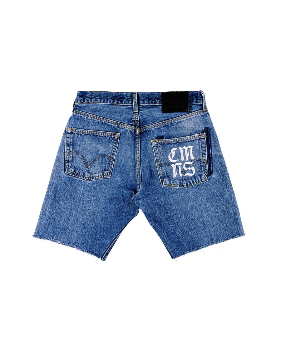 Back view of blue denim shorts with raw hems, two pockets, white lettering on one pocket, and a black waistband patch.