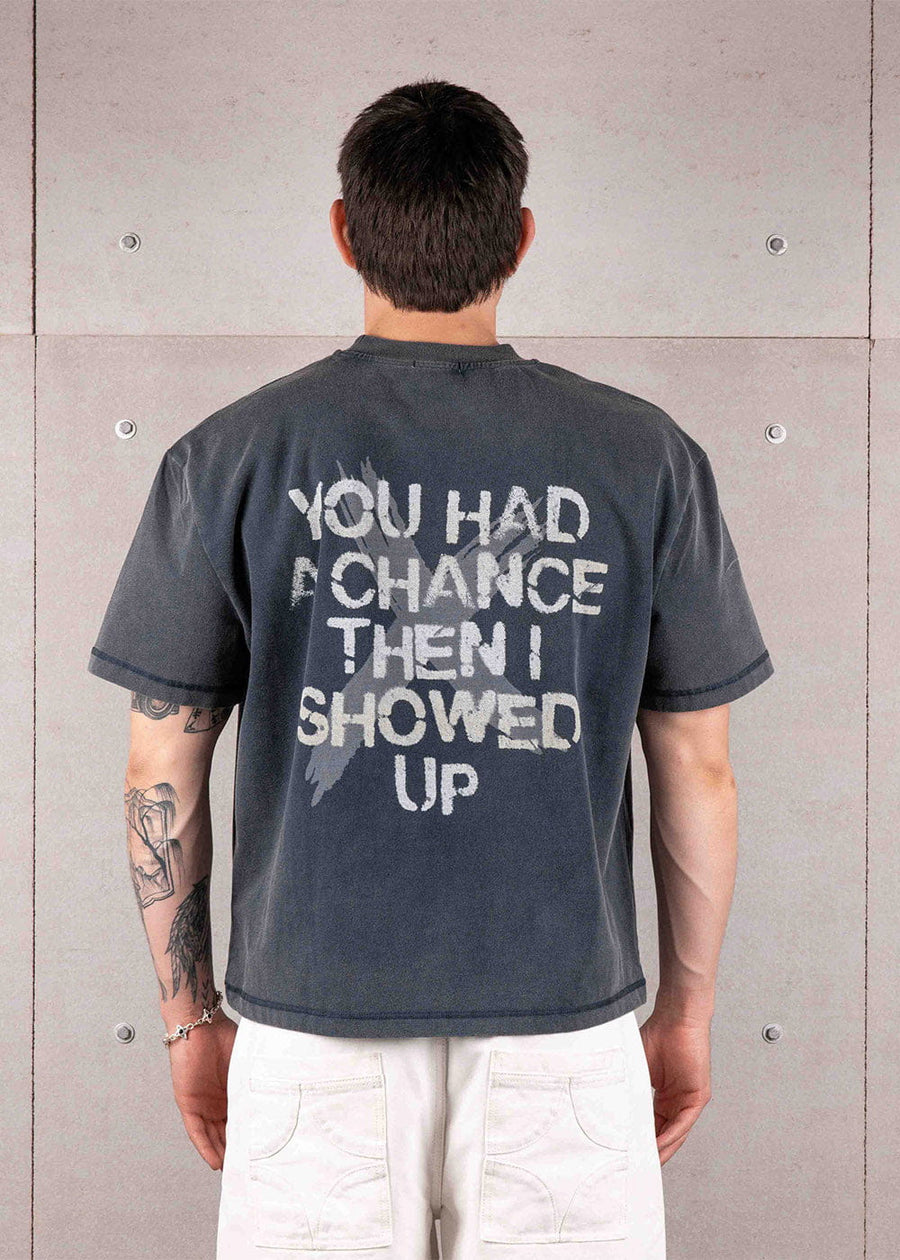 Back view of a person wearing a faded navy T-shirt with large white slogan lettering and white pants against a concrete wall.