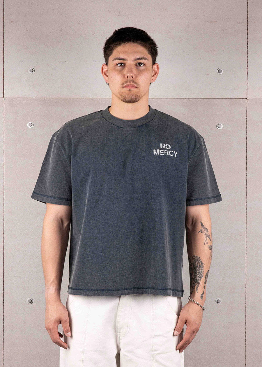 Man wearing a faded navy crewneck T-shirt with white NO MERCY chest text and white pants against a concrete wall.