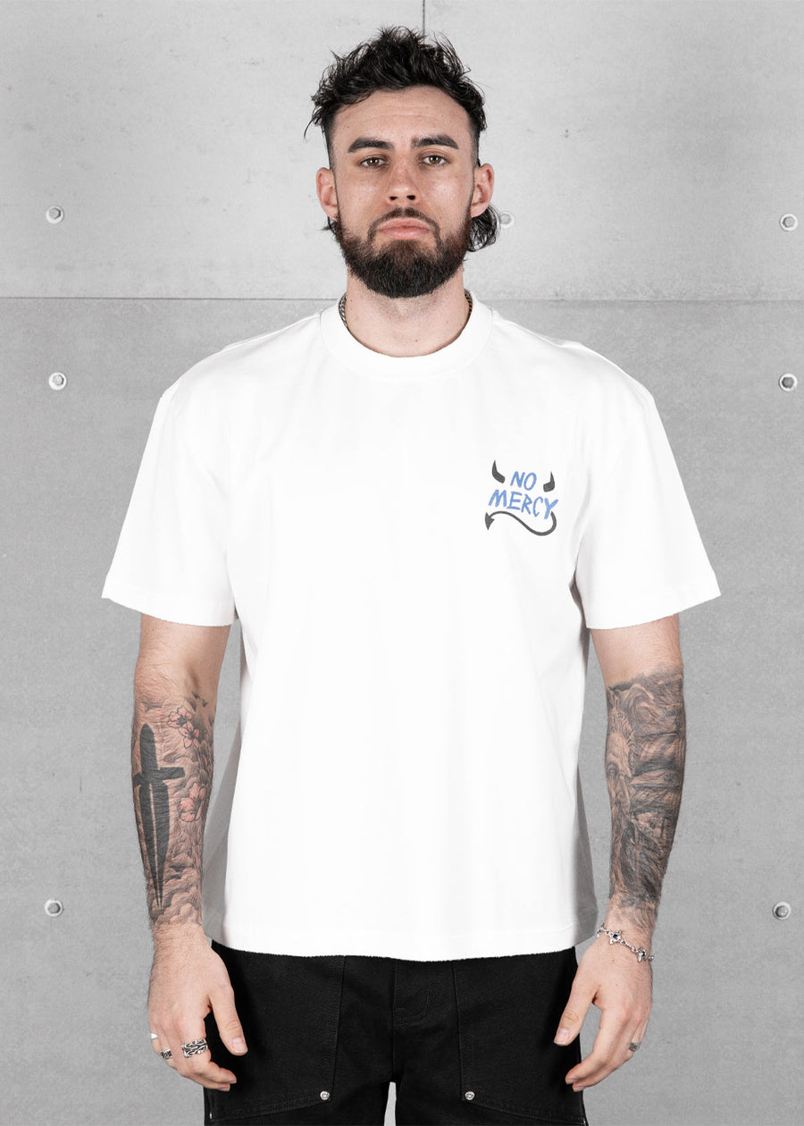 Man wearing a white short-sleeve T-shirt with a blue and black No Mercy chest graphic against a gray concrete wall.