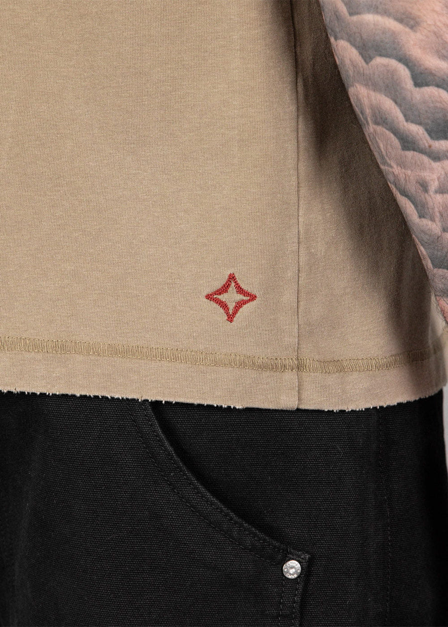 Close-up of a tan knit shirt with a raw hem and red embroidered star, worn over black pants beside a tattooed arm.