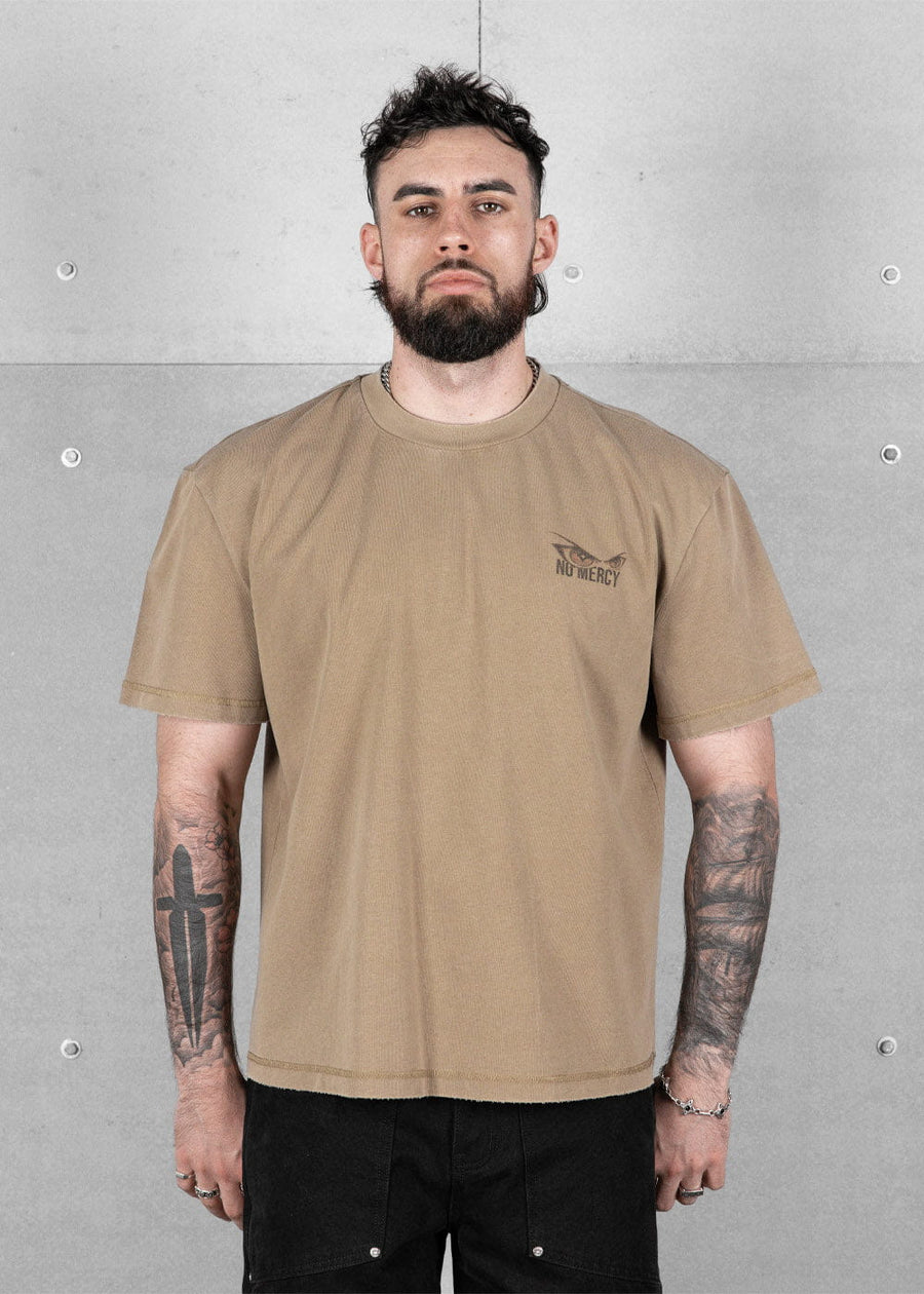 Man wearing a tan crewneck T-shirt with a small No Mercy chest logo and black pants against a gray concrete wall.