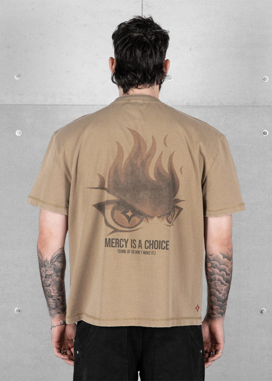 Back view of a model wearing a tan T-shirt with a flaming eye graphic and MERCY IS A CHOICE text against a gray wall.