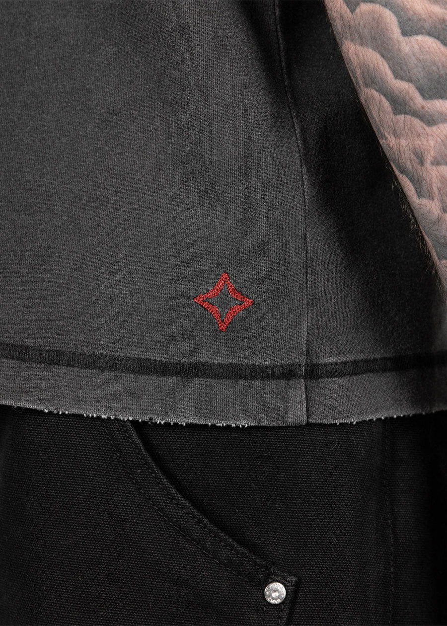 Close-up of a charcoal knit shirt with a red embroidered diamond and raw hem, worn over black jeans beside a tattooed arm.