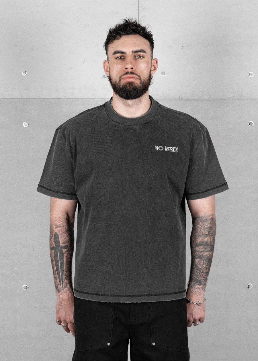 Man wearing a washed charcoal crewneck T-shirt with a small No Mercy chest logo against a gray concrete wall.