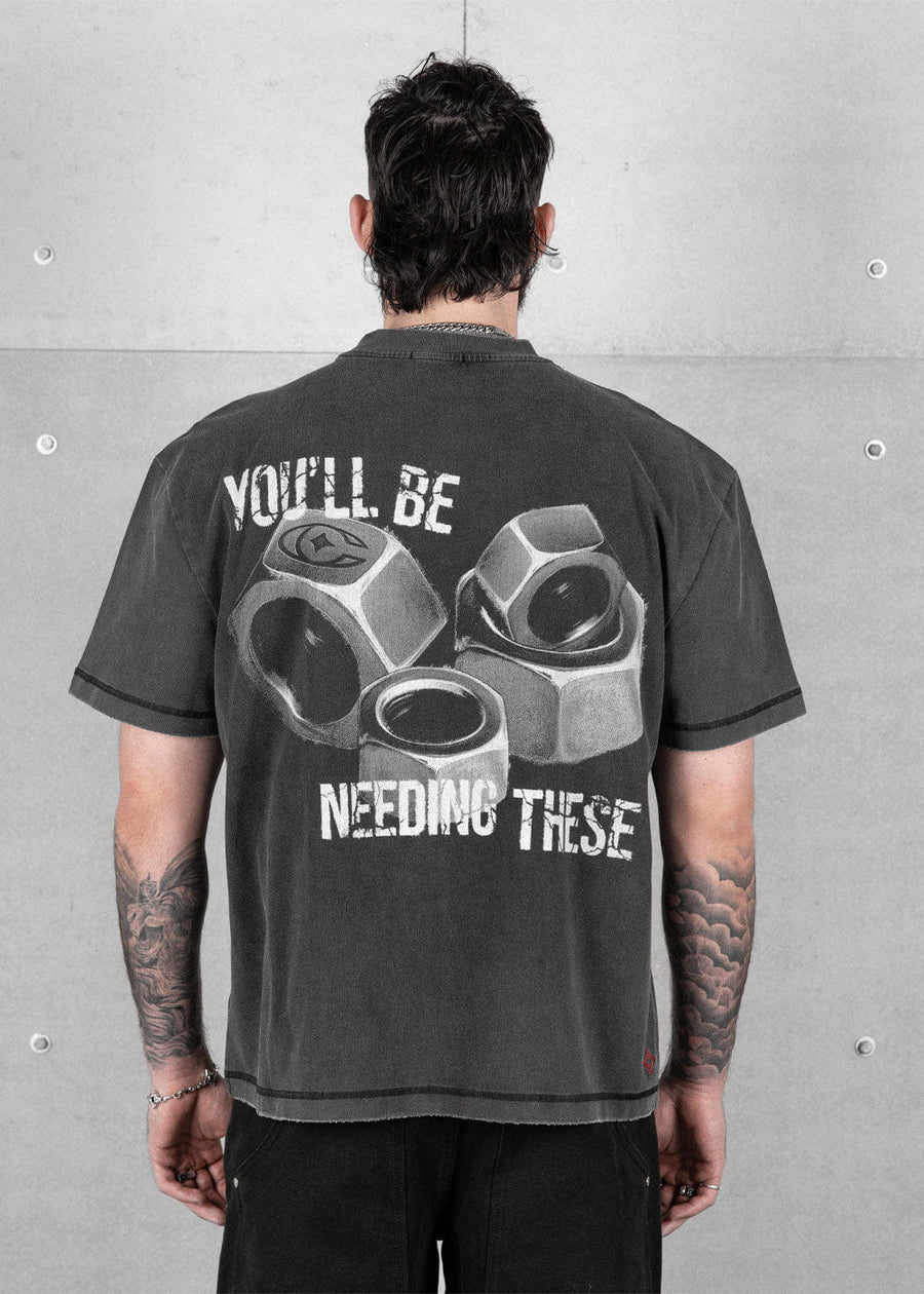 Back view of a model wearing a charcoal T-shirt with large hex nut graphics and white lettering against a concrete wall.