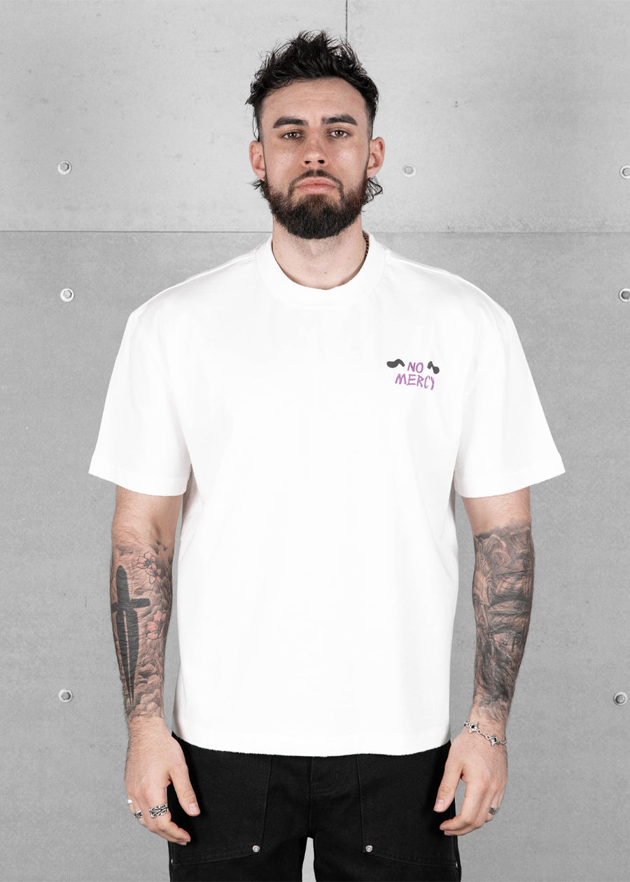 Man wearing a white T-shirt with a small purple NO MERCY chest graphic and black pants against a gray concrete wall.