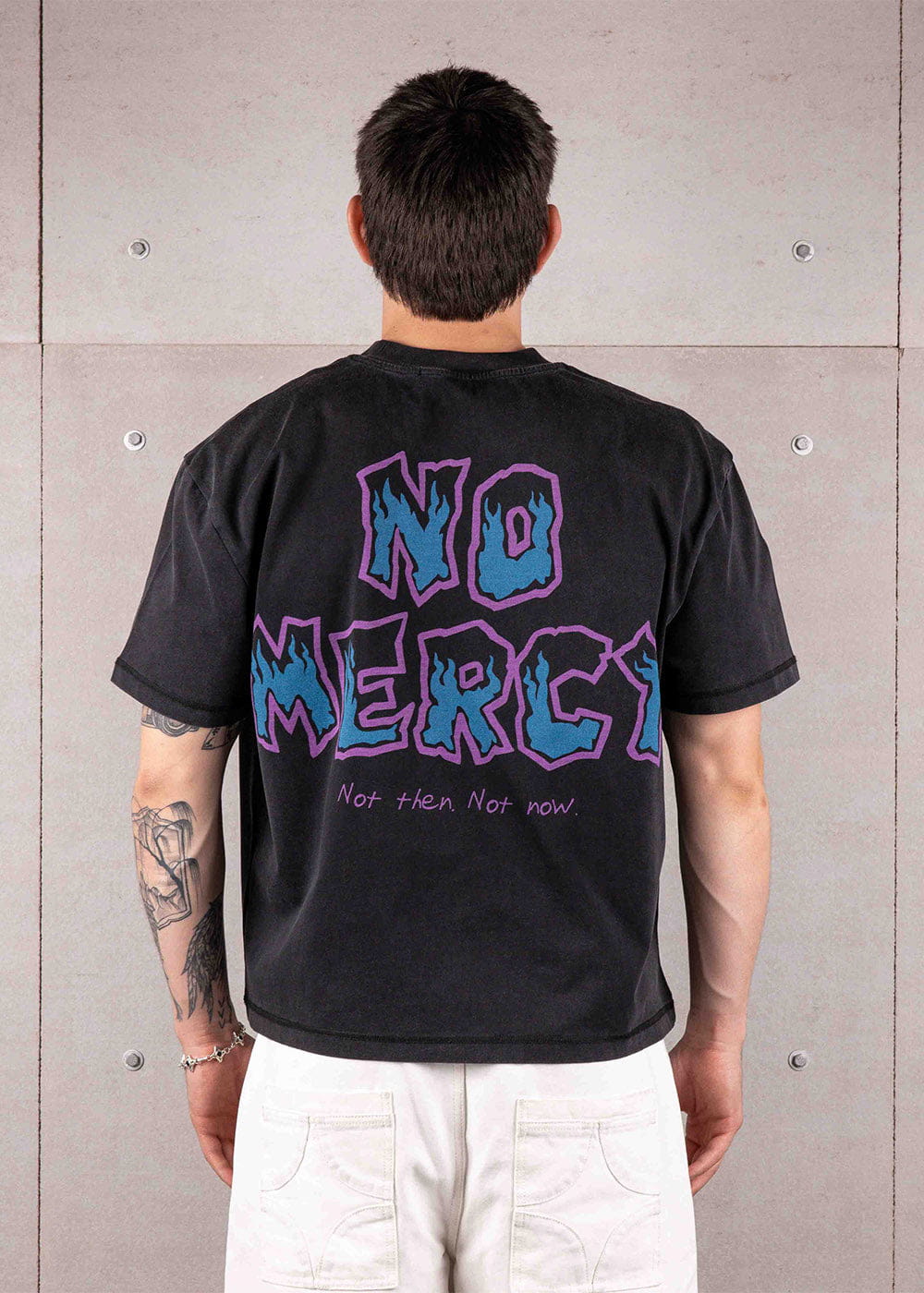 Back view of a person wearing a black T-shirt with blue and purple NO MERCY graphics and white pants against a concrete wall.