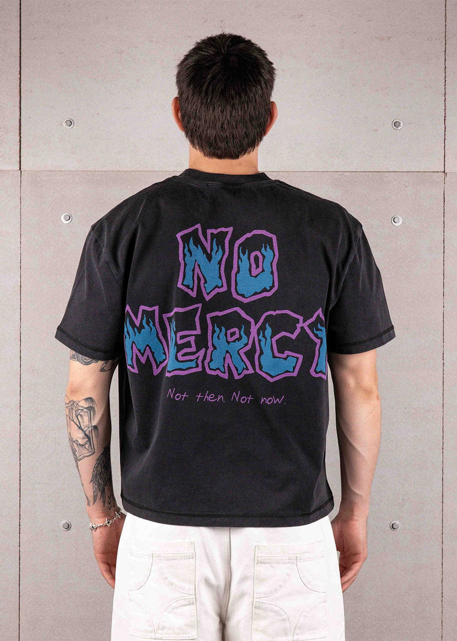 Back view of a person wearing a black T-shirt with blue and purple NO MERCY graphics and white pants against a concrete wall.