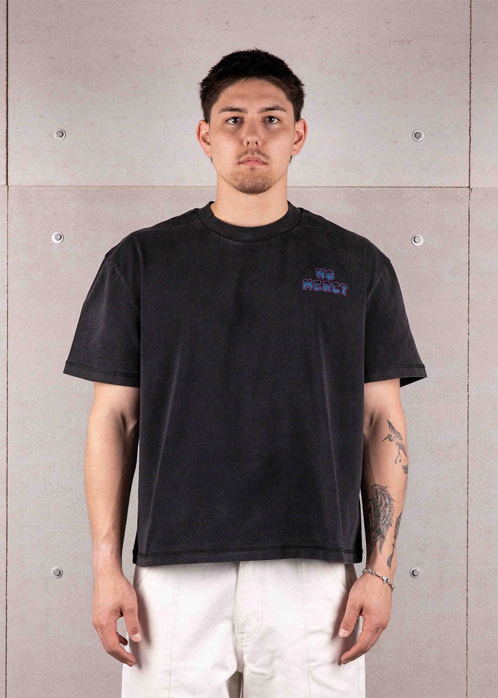 Man wearing a washed black crewneck T-shirt with small No Mercy chest lettering and white pants against a concrete wall.