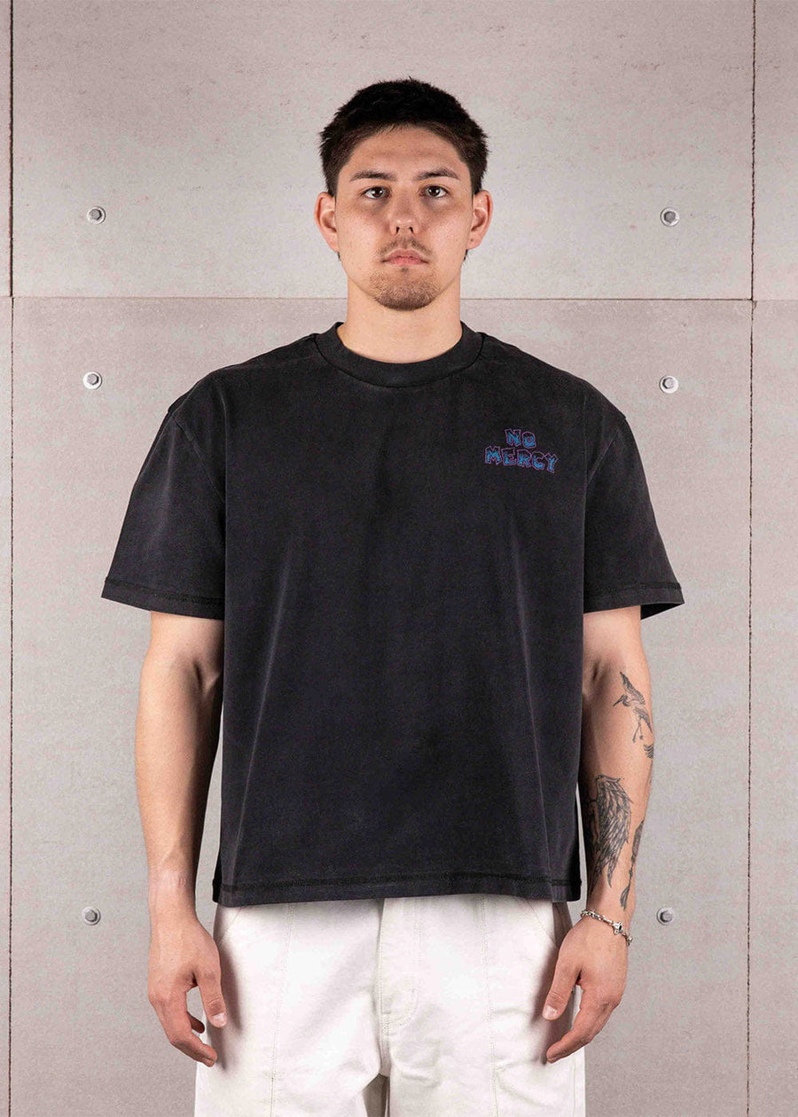 Man wearing a washed black crewneck T-shirt with small No Mercy chest lettering and white pants against a concrete wall.
