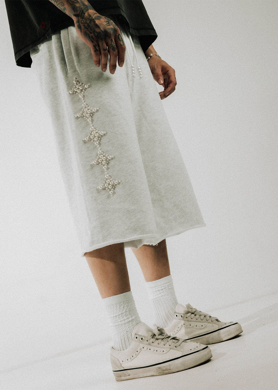 Model wearing light gray wide-leg shorts with pearl-beaded motifs, a black shirt, white socks, and cream sneakers.