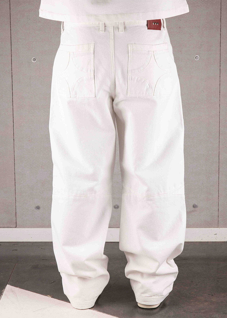 Back view of white wide-leg pants with star-stitched rear pockets and a red waistband patch, worn against a gray wall.