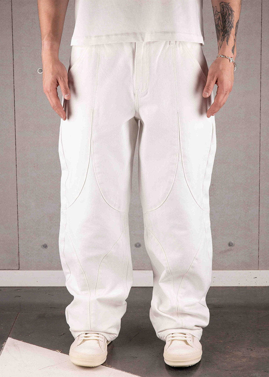 Model wearing white wide-leg pants with curved panel seams and white sneakers against a gray concrete wall.